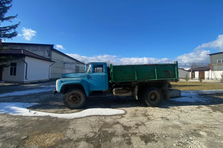 Un camion basculant ZIL vechi, albastru și verde, în curtea cramei, sub un cer senin, cu urme de zăpadă