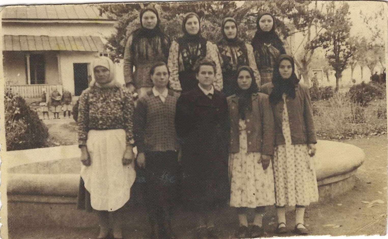Fotografie de epocă: un grup de femei din Răzeni, cele mai multe cu basmale, în fața fântânii satului; în spate, o casă cu prispă